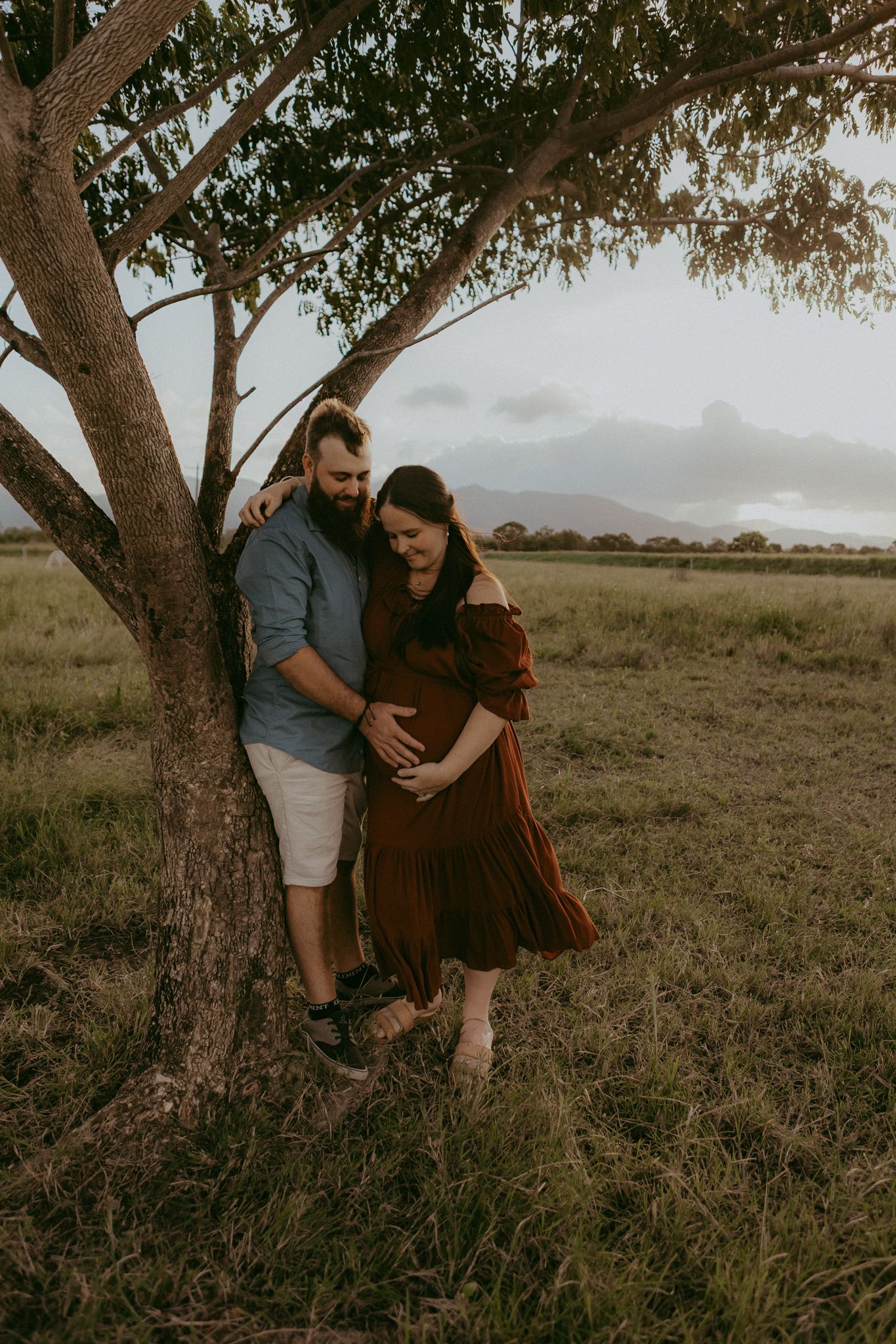 Couple leaning against a tree in a grassy field, cradling pregnant belly during golden hour maternity shoot.