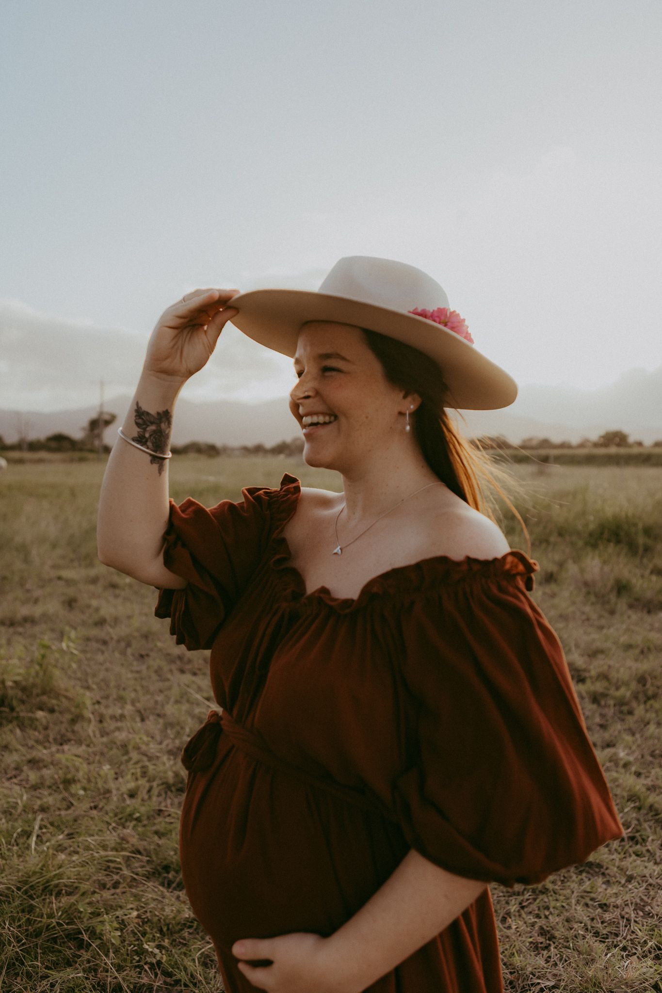 Laughing pregnant woman in rust off-shoulder dress holding a white hat in an open field at sunset.