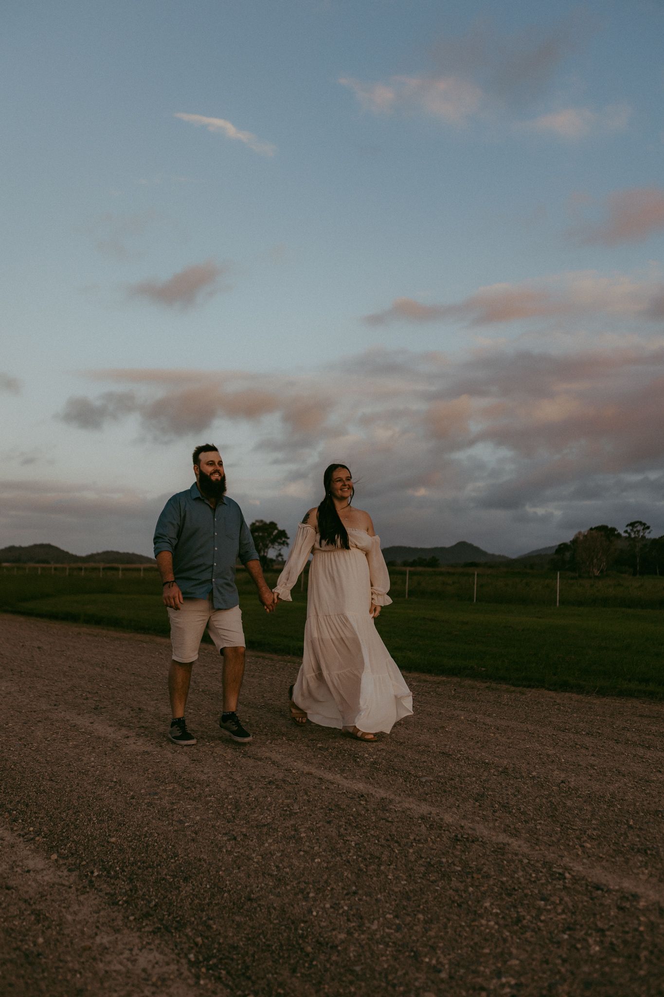 Pregnant couple holding hands walking on a gravel road under a pastel sunset sky.