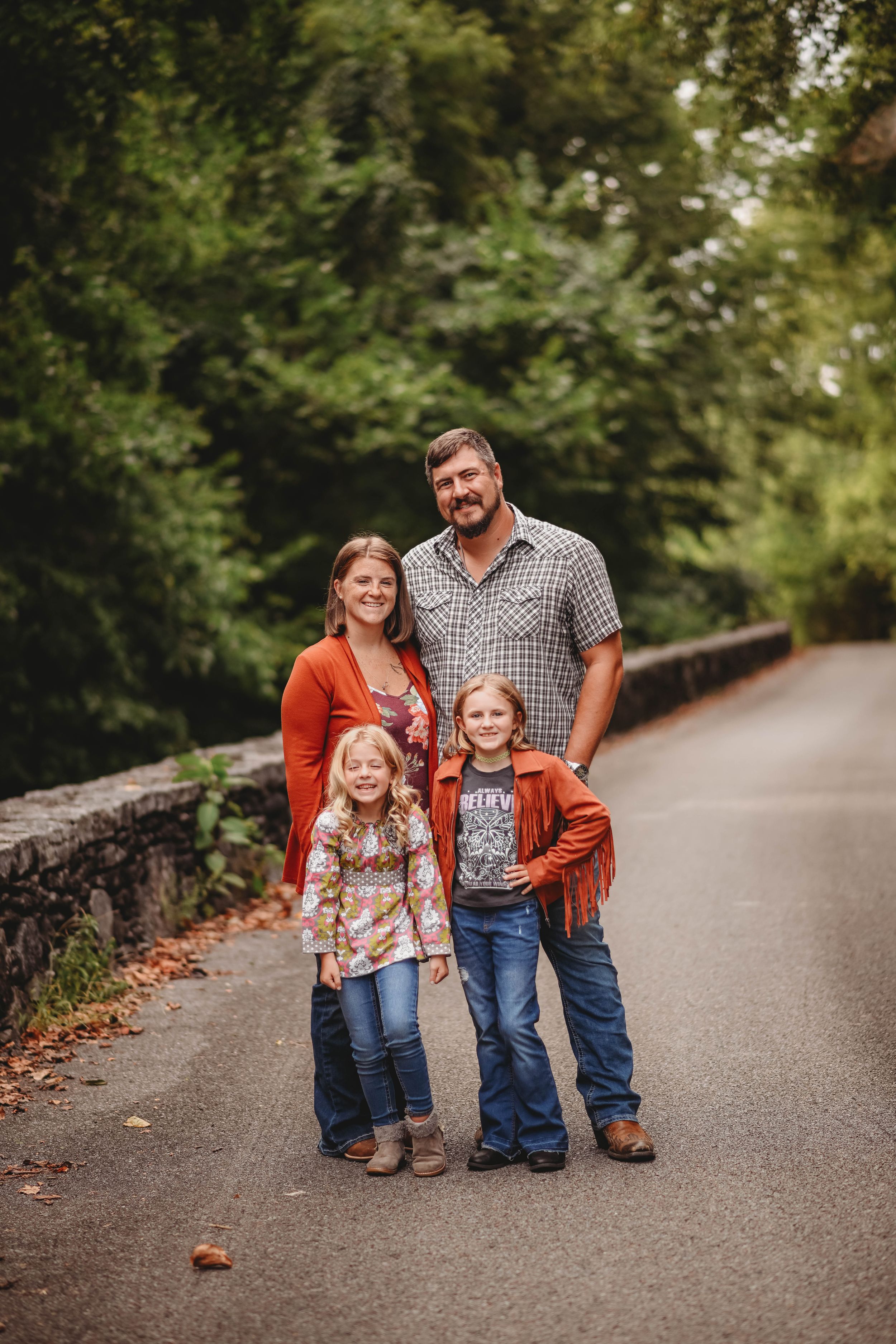 A family of four standing together on a stone-lined pathway in an autumn setting.