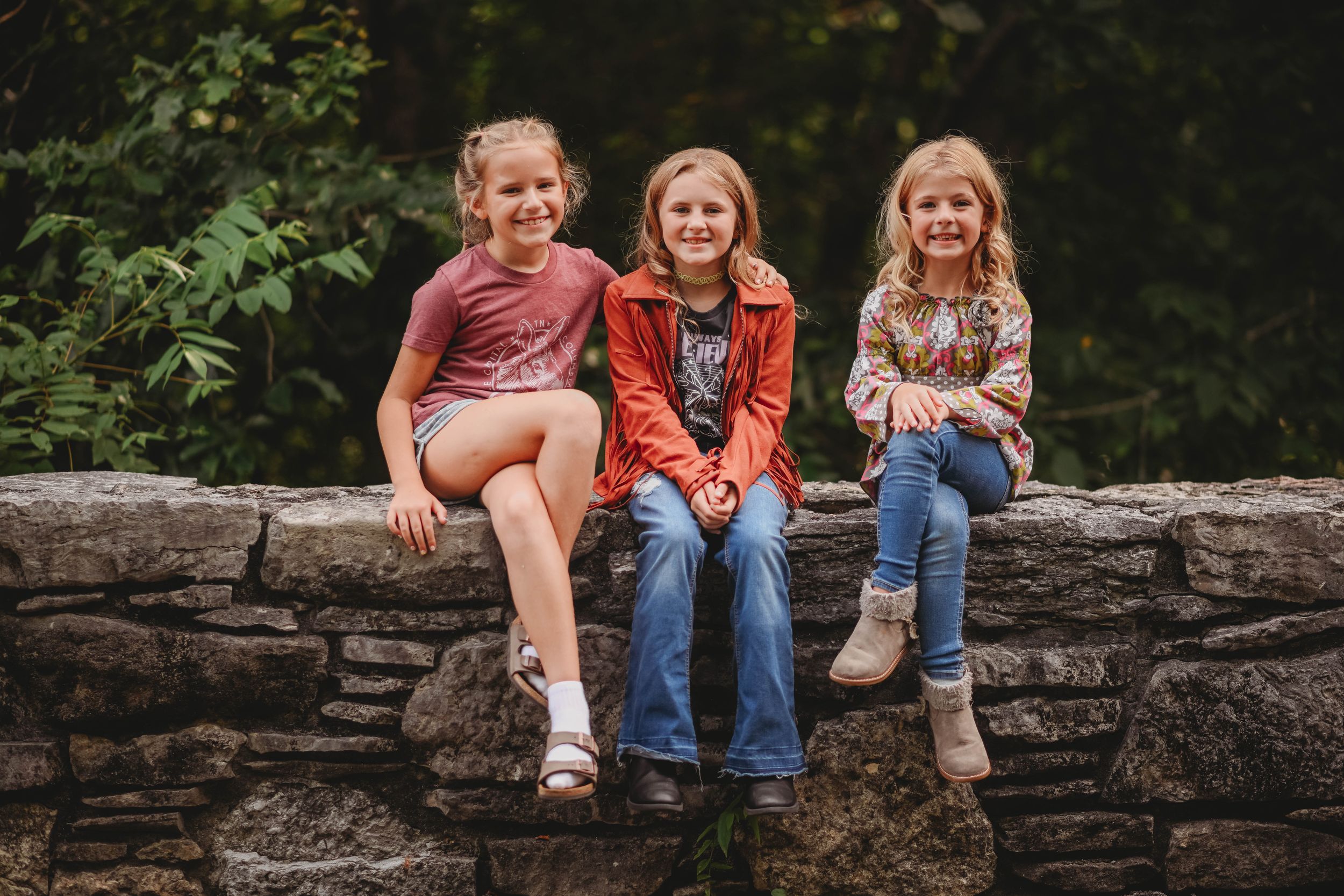 Three children sitting casually on a stone wall outdoors.
