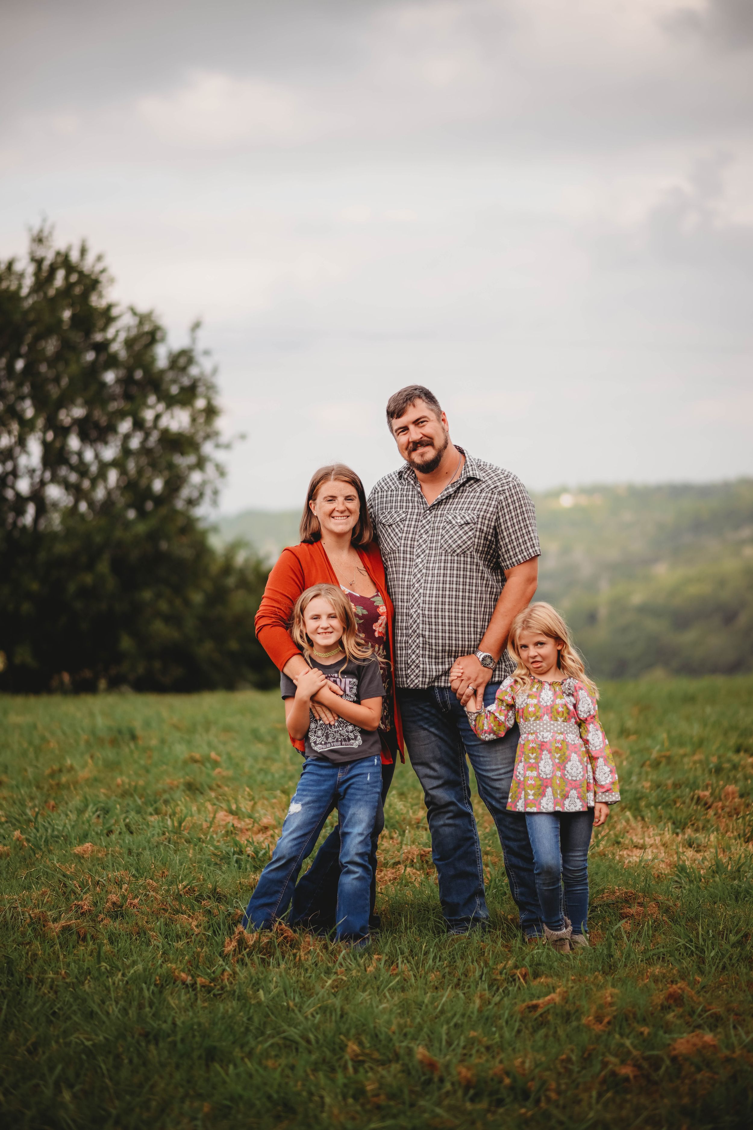 A family of four posing together in a grassy field with rolling hills in the background.