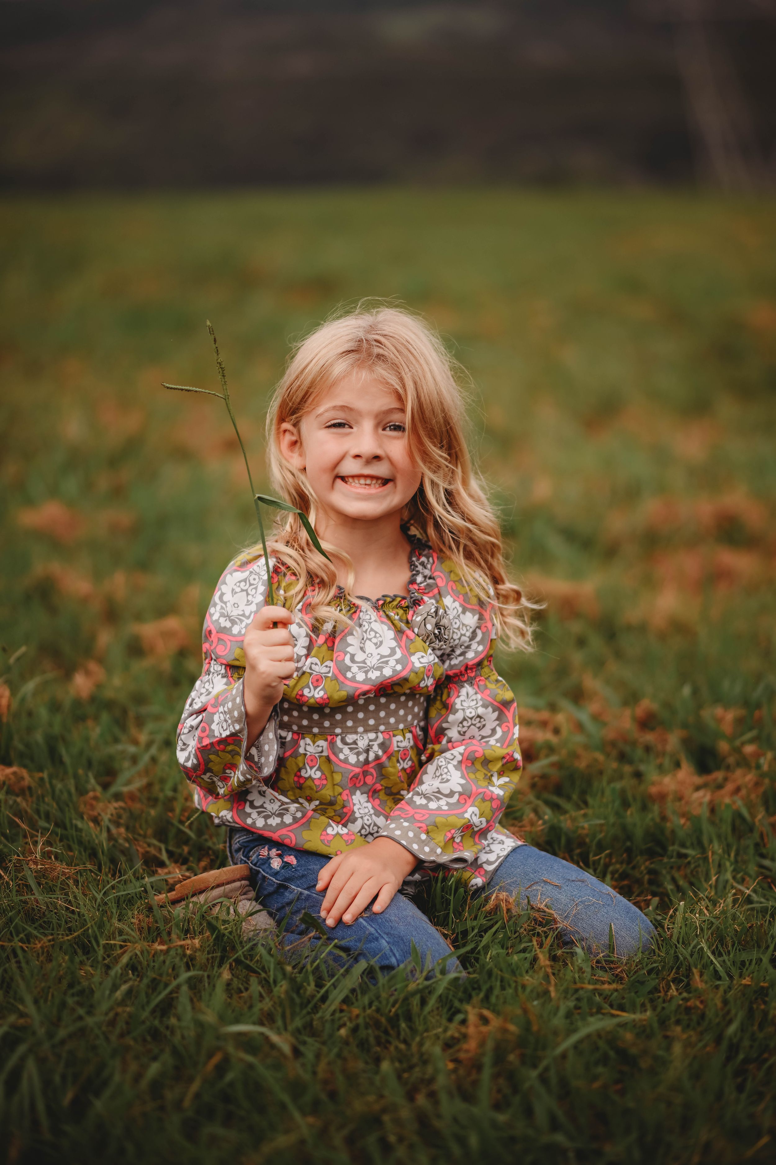 A child sitting in grass holding dried plants and smiling brightly.