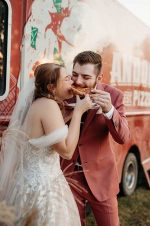 A joyful couple in wedding attire share a pizza slice together in front of a red food truck.
