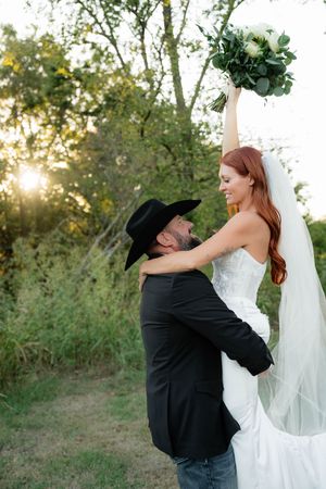 Groom in black cowboy hat lifts bride in white dress as she raises bouquet, golden sunset in background.