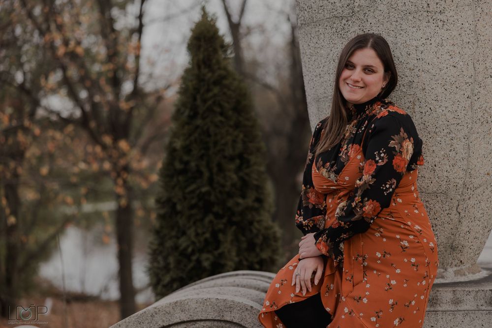 Woman in bright orange coat and floral scarf poses against stone wall backdrop during autumn photoshoot.