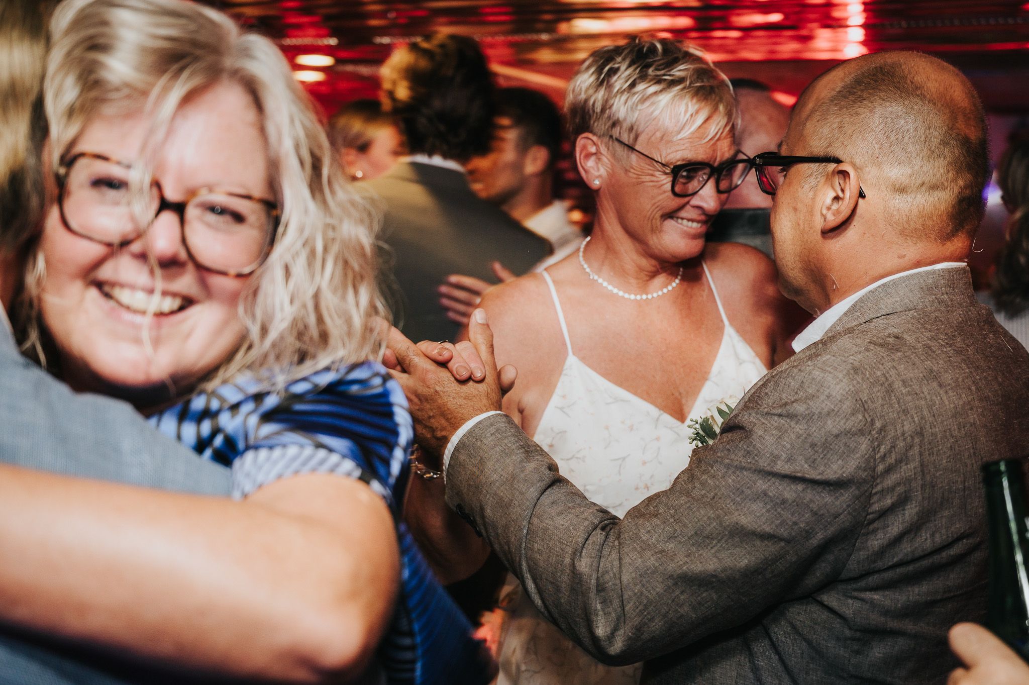 People dancing and celebrating at a lively wedding reception with red ambient lighting.