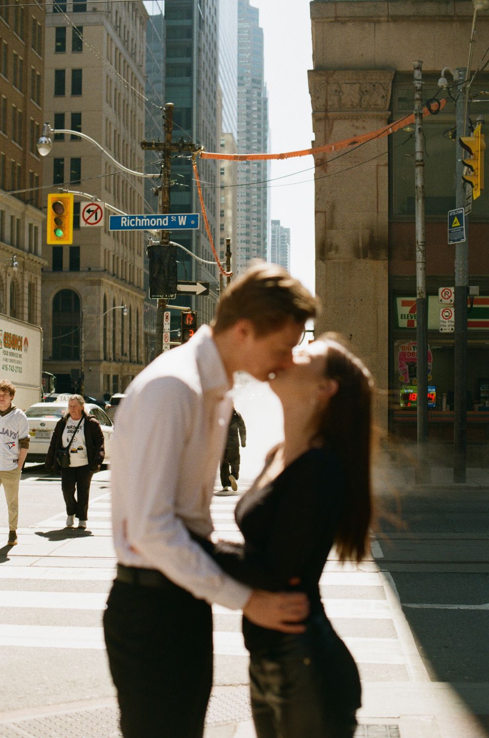 Couple sharing a romantic kiss on Richmond Street in downtown Toronto, with city buildings and pedestrians in the background.