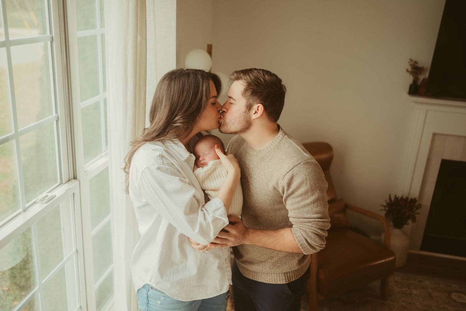 New mom and dad kiss in living room in front of window with baby on mom's chest, during lifestyle newborn session in Clarksville, Tennessee