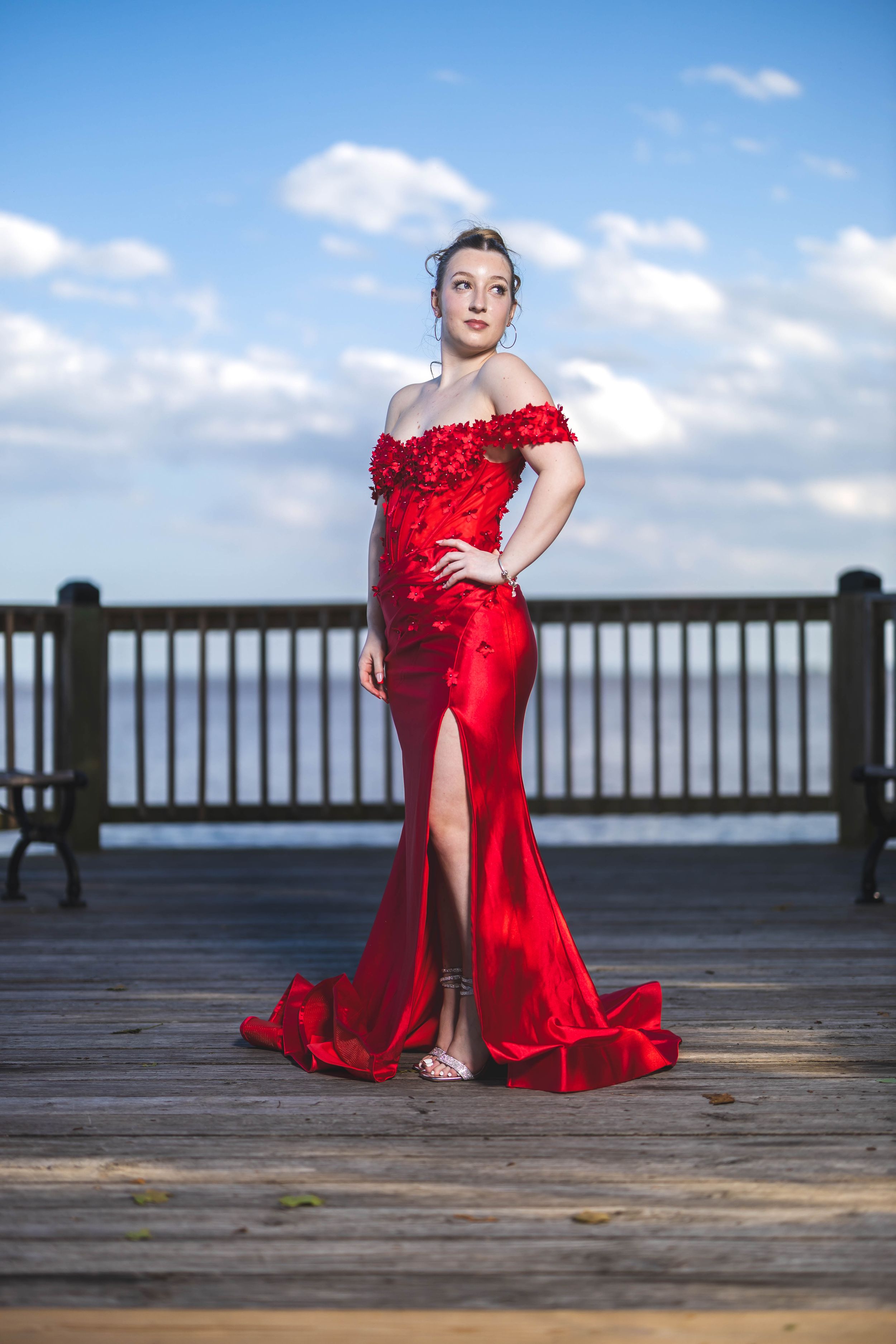 A person in a flowing red formal gown poses gracefully on a wooden boardwalk against a bright blue sky.