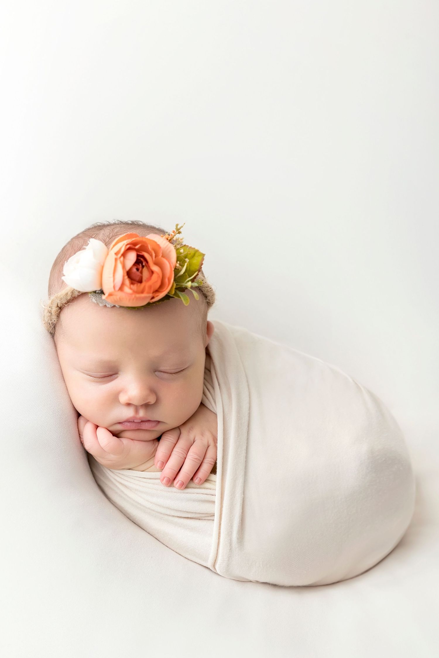 Sleeping newborn wrapped in cream fabric wearing delicate peach floral headpiece.