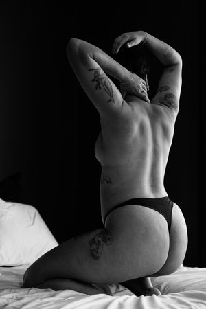 Black and white boudoir photo of tattooed woman kneeling on bed, showcasing muscular back and tattoos.