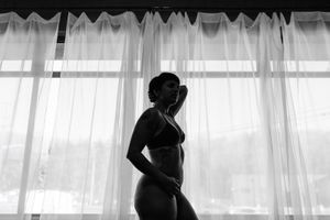 Black and white silhouette of woman in lingerie posing by sheer curtained window in boudoir photo.