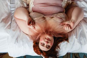 Overhead view of a plus-size redhead woman in pink tulle dress lying on white bed with pearl necklace.