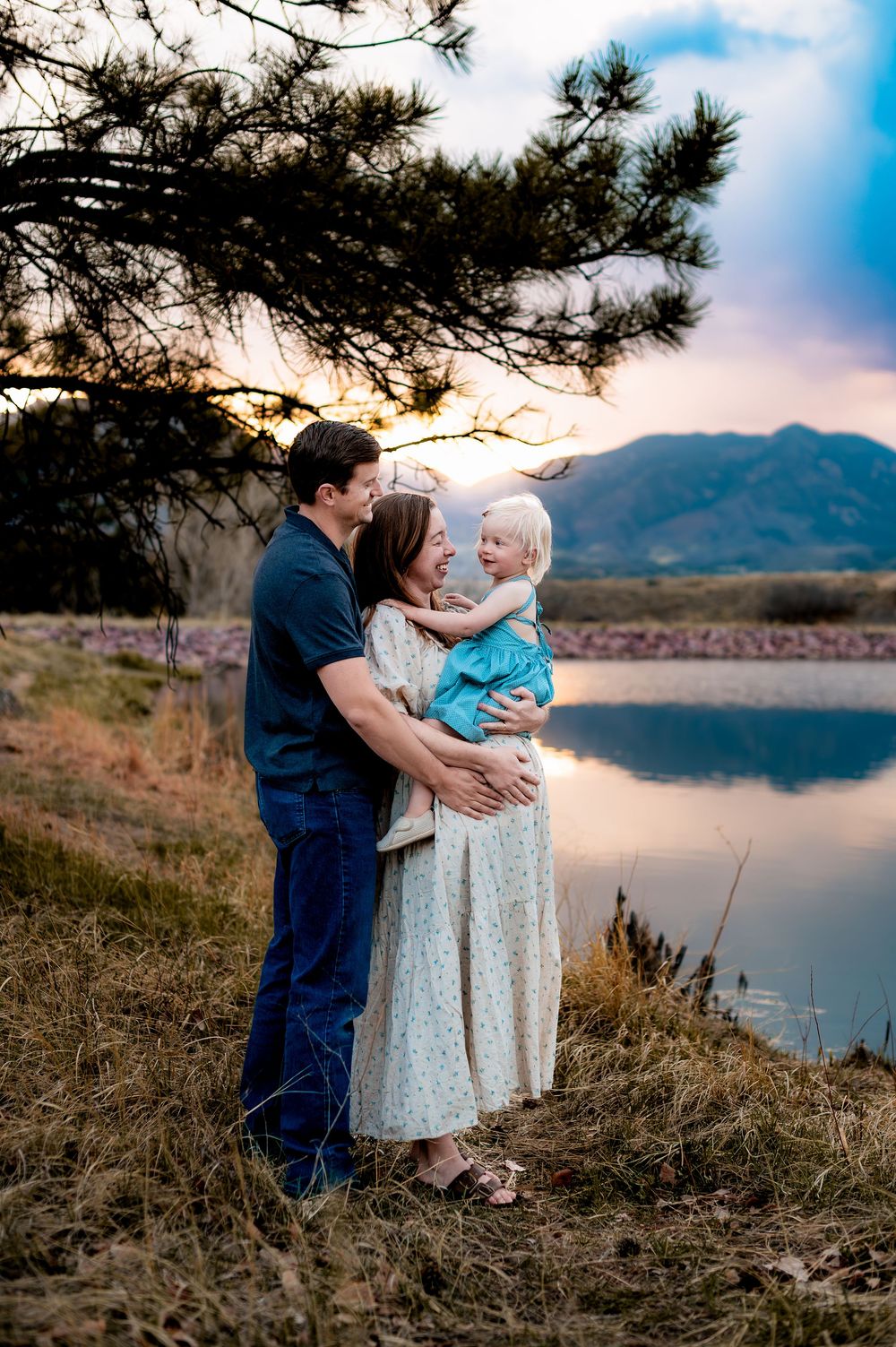 A family shares a tender moment by a mountain lake at sunset, with pine trees framing the scenic backdrop.