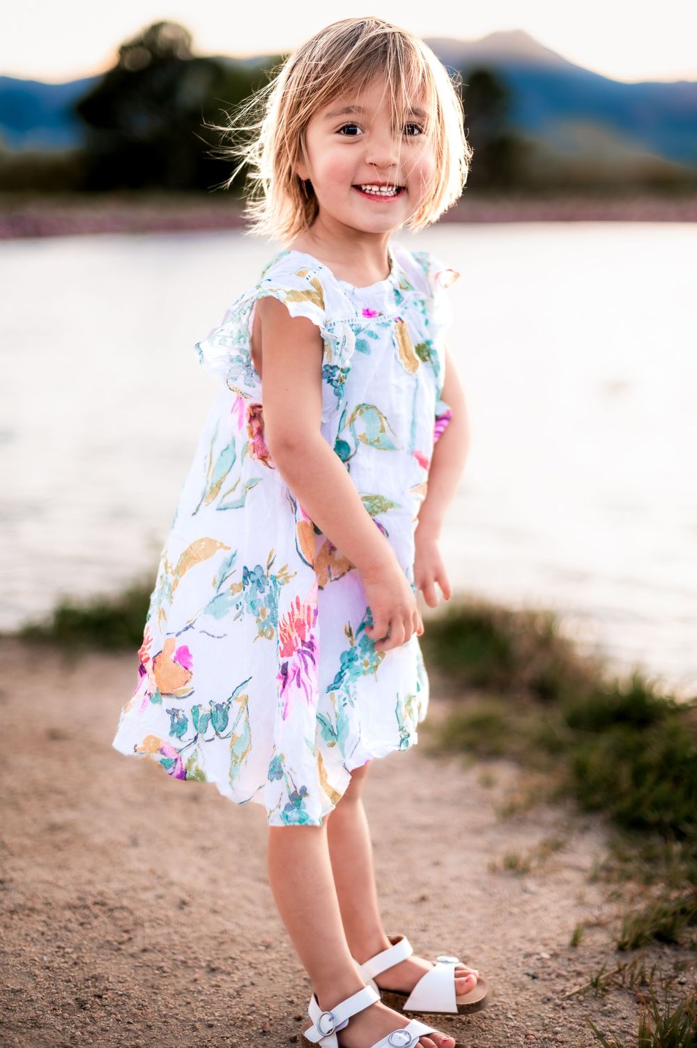 A floral print summer dress and white sandals shown against a mountain backdrop during sunset.