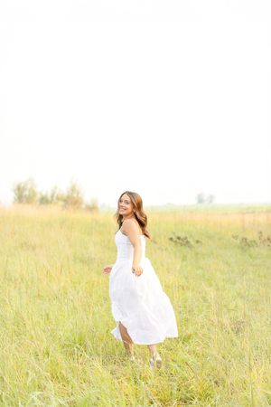 A high school girl in a flowing white dress twirls gracefully through a sun-drenched meadow of tall grass at family land in Clear Lake