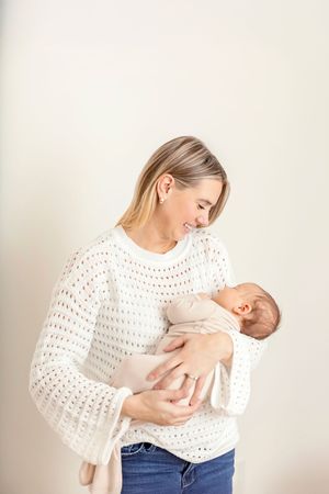 A parent in a white knit sweater and jeans holds and gazes lovingly at their newborn baby against a light background.