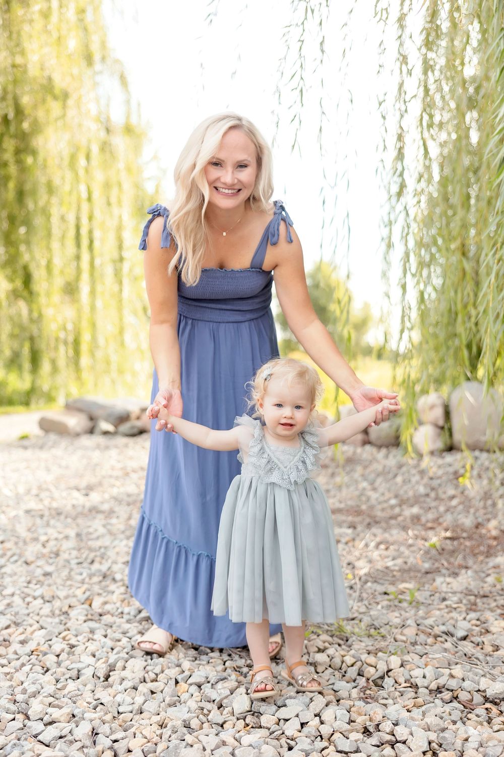 Mother and toddler in matching blue dresses stand together on rocky ground beneath a willow tree.