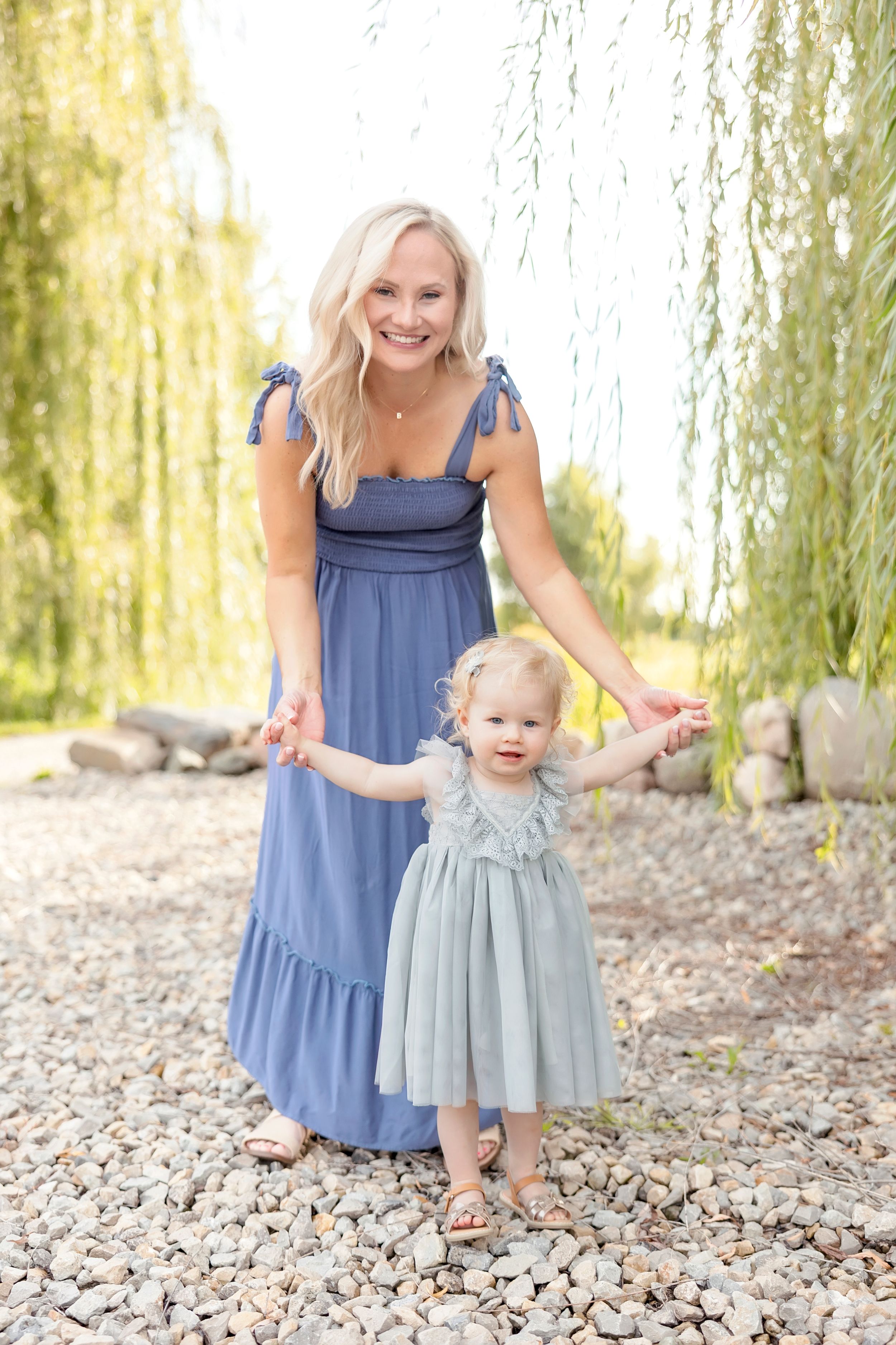 Mother and toddler in matching blue dresses stand together on rocky ground beneath a willow tree.