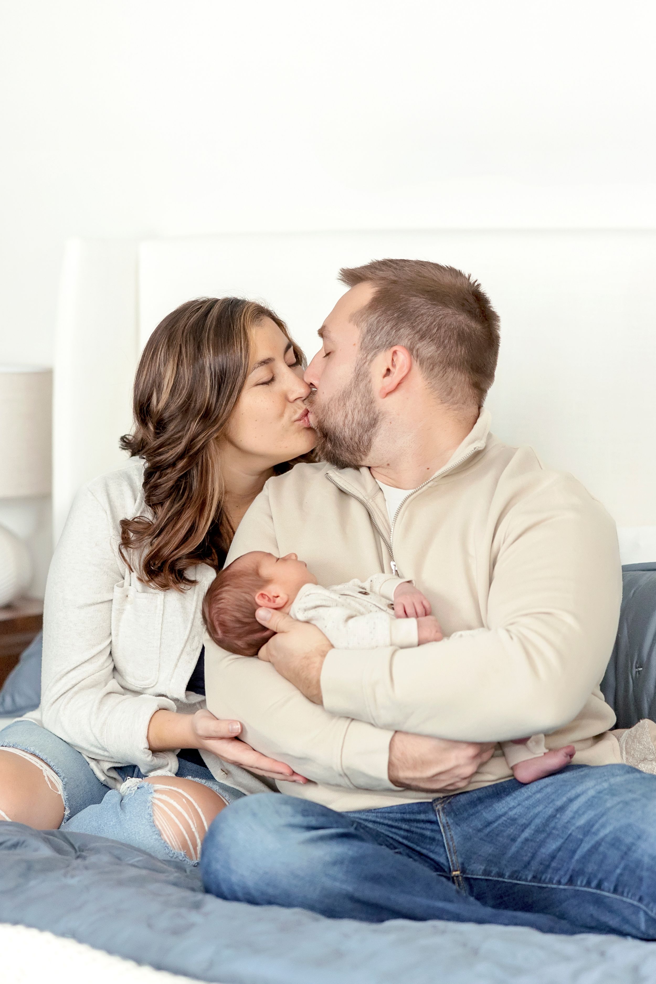 A couple shares a tender kiss while holding their newborn baby on a cozy bed with blue bedding.