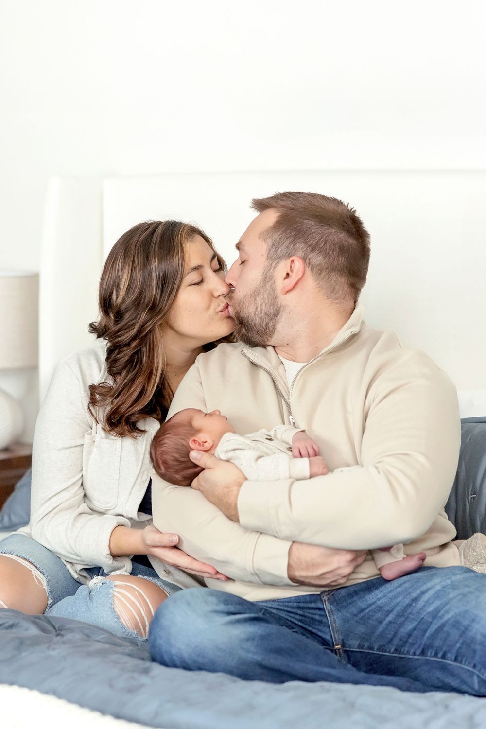 A couple shares a tender kiss while holding their newborn baby on a cozy bed with blue bedding.