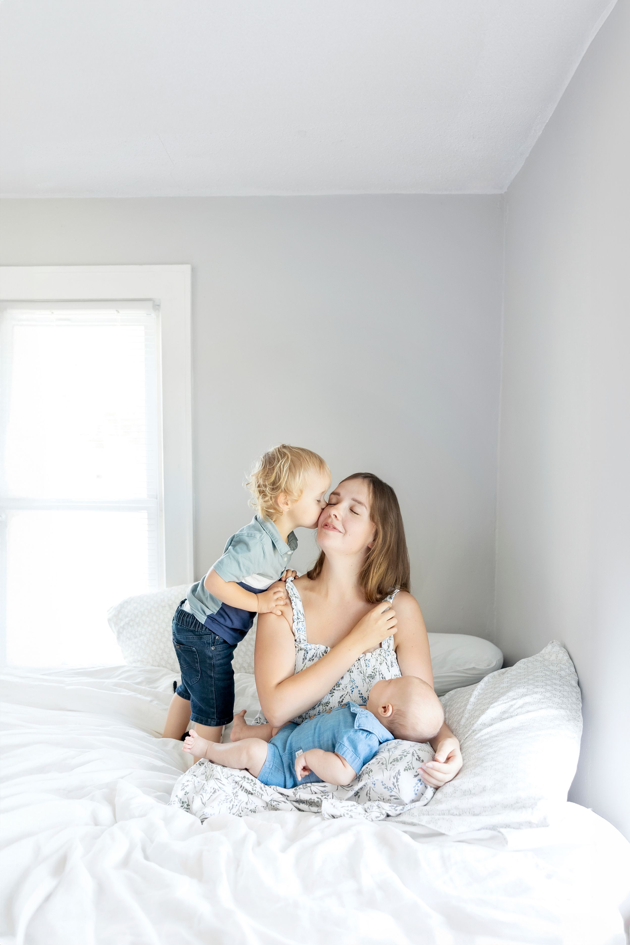 Mother and two children sitting together on a white bed in a bright, minimalist bedroom in Garner Iowa during in home newborn lifestyle
