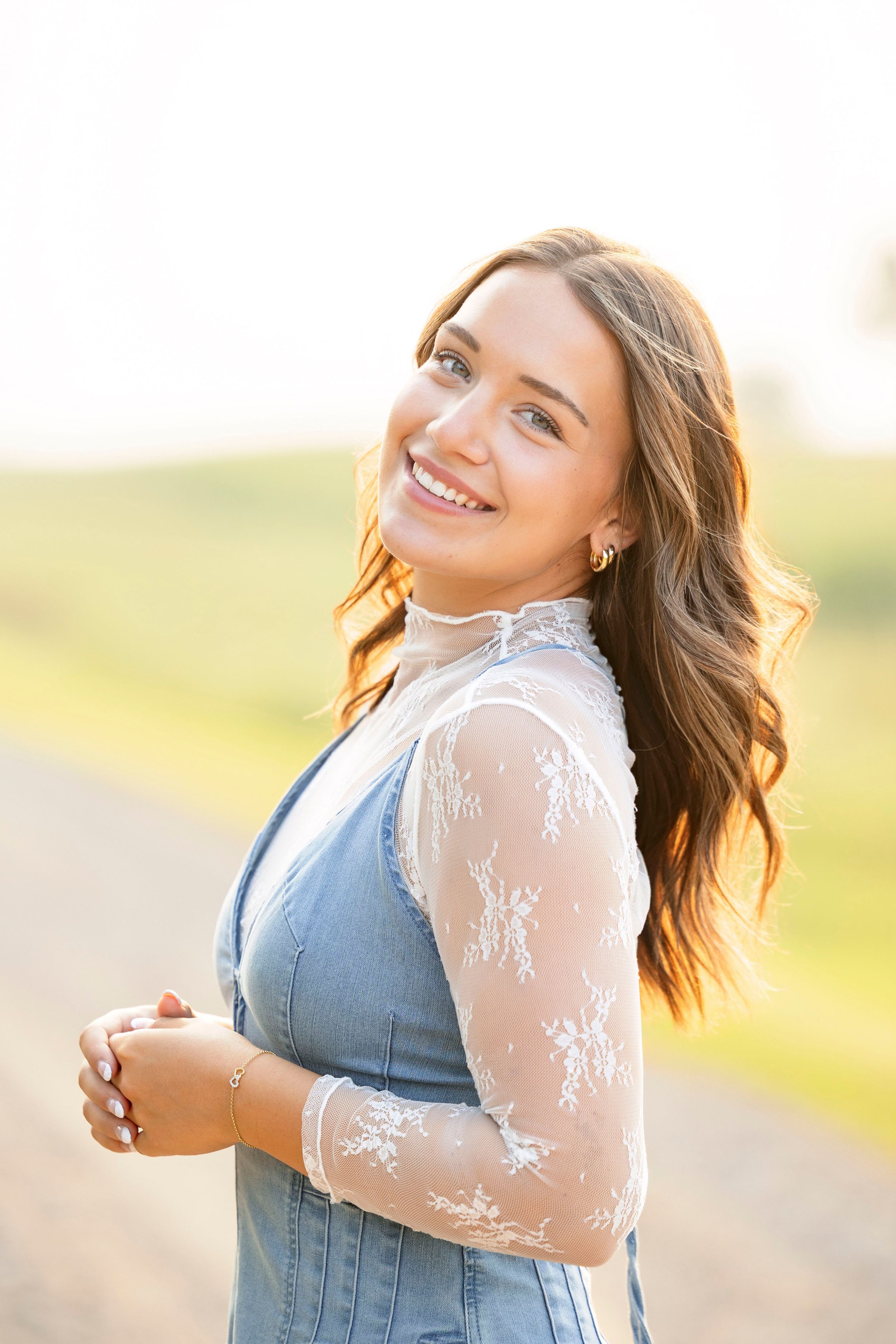 A person wearing a denim dress with sheer lace sleeves poses outdoors in warm sunlight with a bright smile.