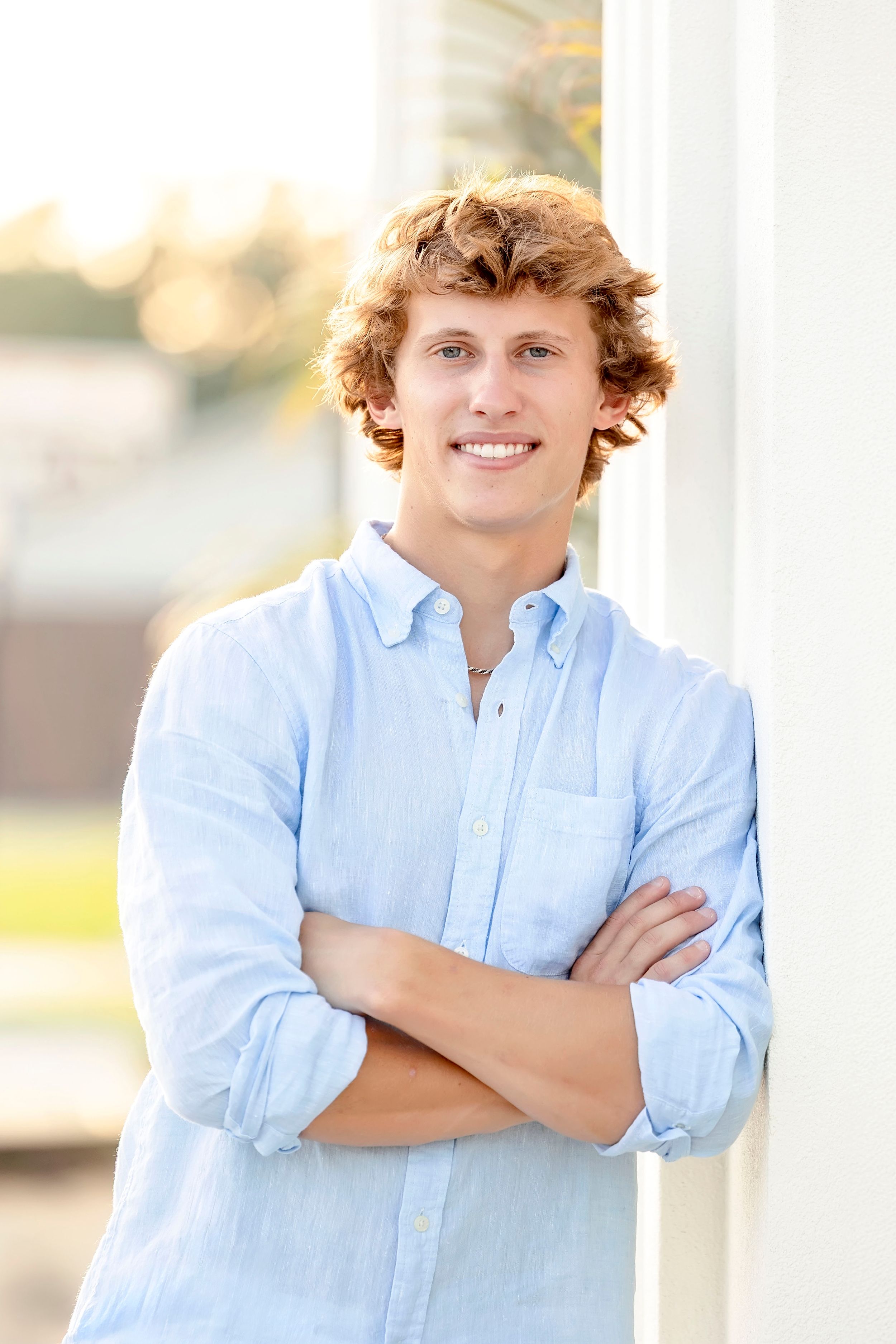 Young person in light blue button-down shirt stands with arms crossed against white pillar outdoors in natural lighting.