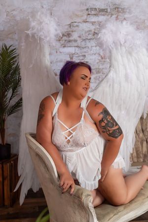 Person in white lingerie posing with angel wings against a rustic backdrop.