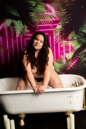 Artistic boudoir photoshoot setup with vintage bathtub, neon pink lights, and tropical palm leaves in the background.