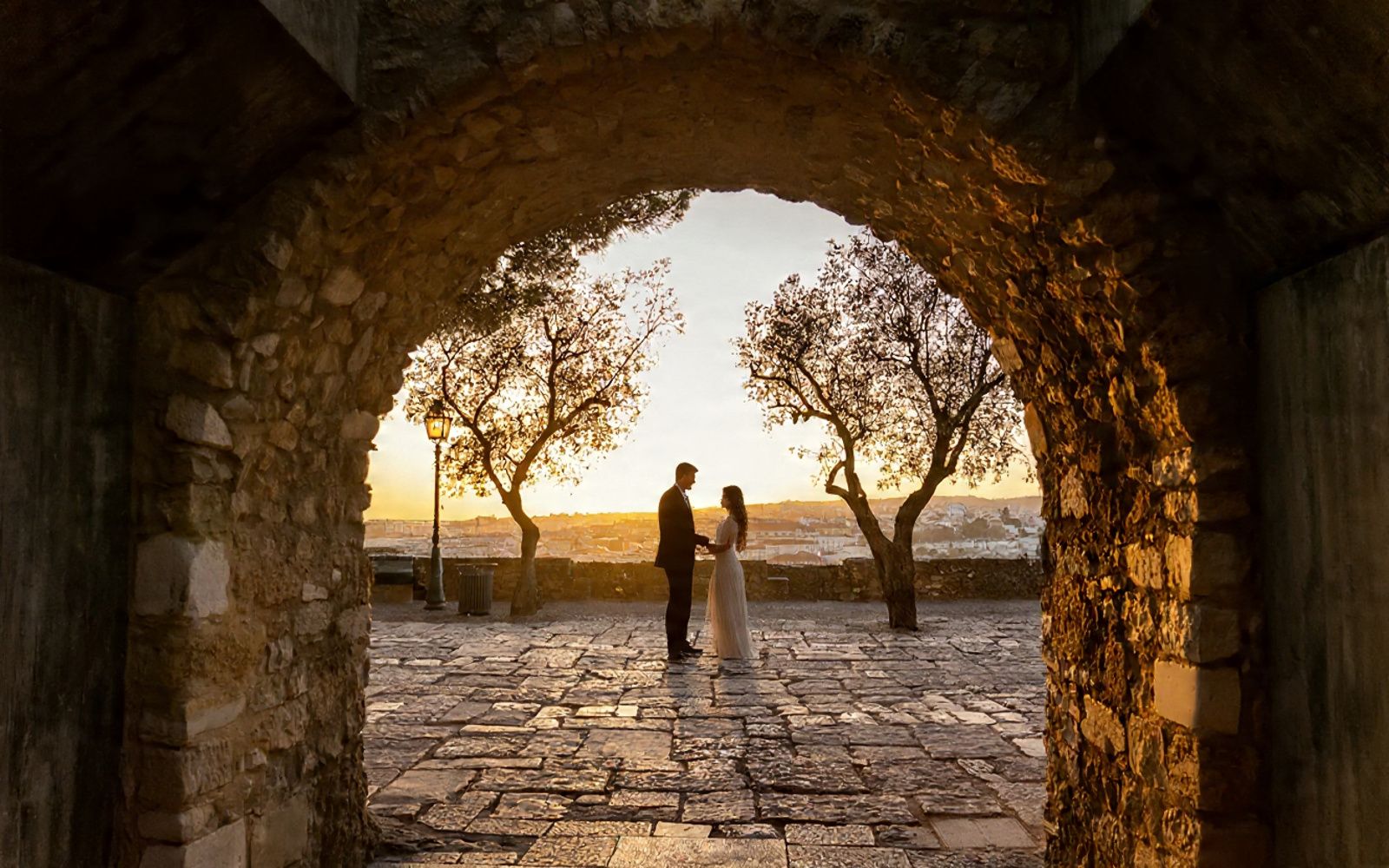 Stone archway frames silhouettes of two people walking against a golden sunset on a cobblestone path.