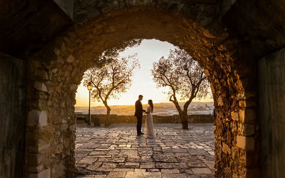 Stone archway frames silhouettes of two people walking against a golden sunset on a cobblestone path.