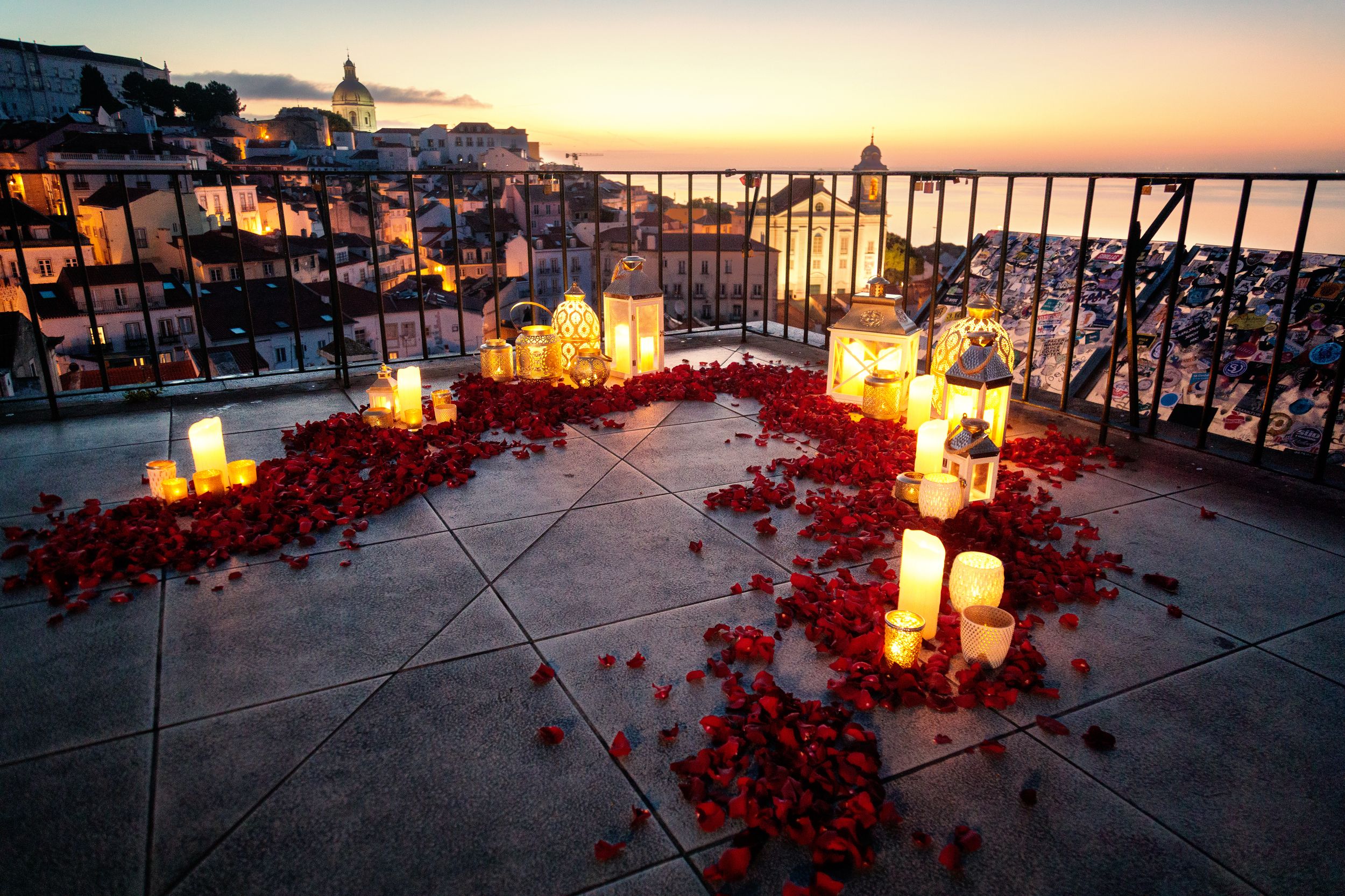 Romantic sunset balcony decorated with red rose petals and glowing candles overlooking the ocean at dusk.