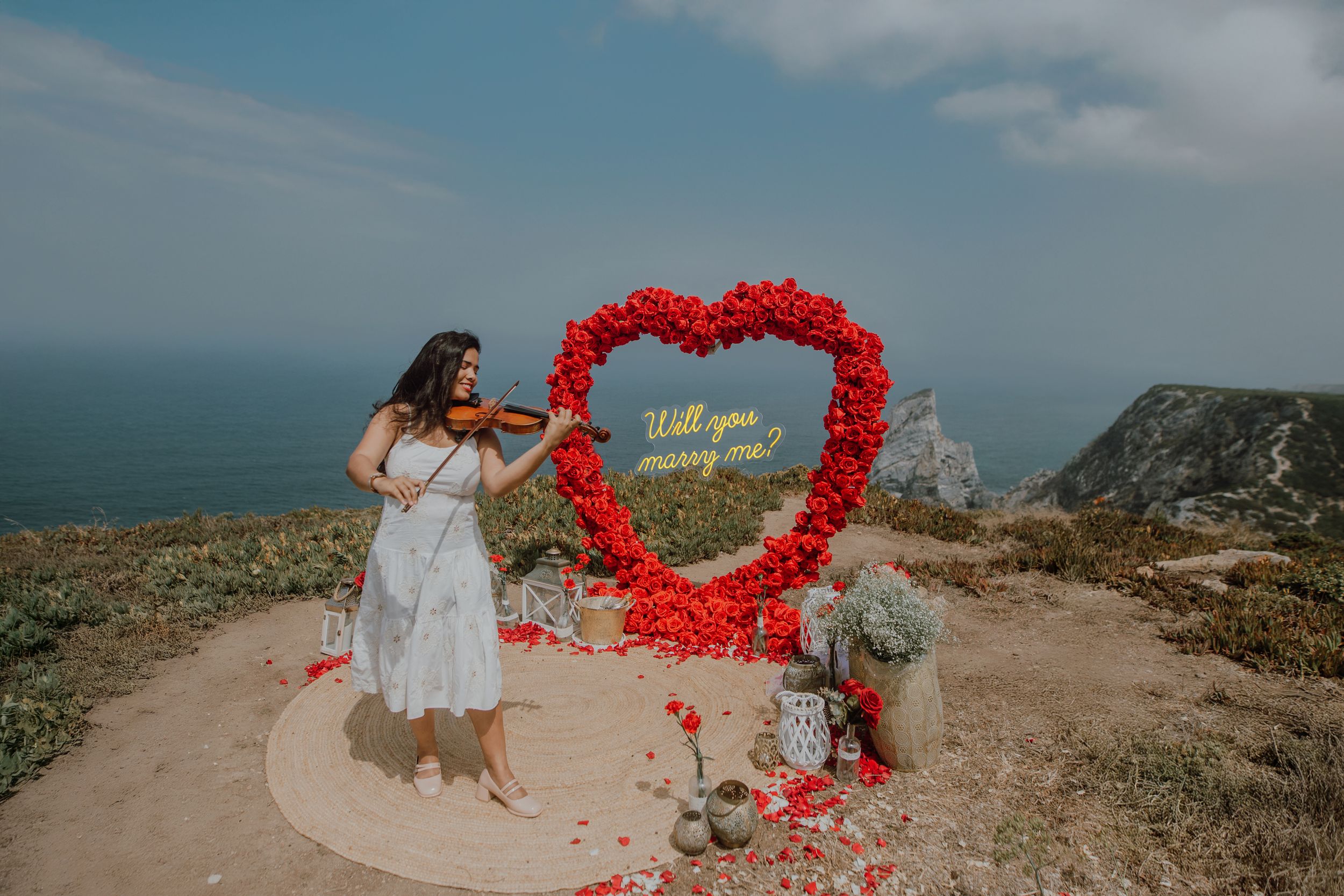 Romantic coastal proposal setup with red rose petals arranged in heart shape on cliff overlooking ocean at sunset.