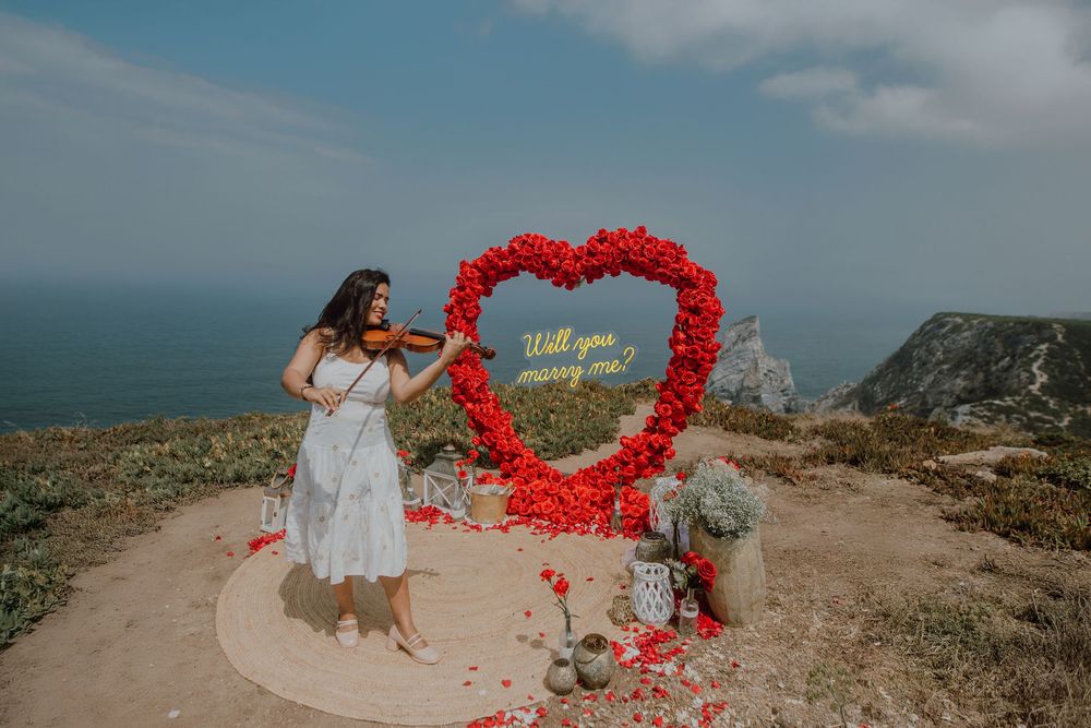 Romantic coastal proposal setup with red rose petals arranged in heart shape on cliff overlooking ocean at sunset.