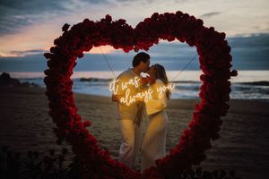 A romantic beach scene with a heart-shaped red rose arch and glowing neon sign at sunset.