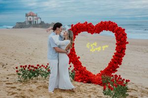 A couple embraces on a sandy beach next to a large red heart-shaped floral arch with a church in the background.