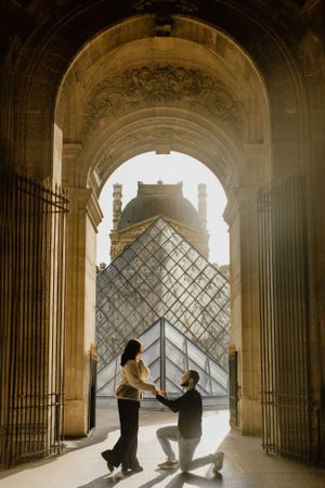 Romantic proposal silhouette under ornate archway at the Louvre Museum with glass pyramid in background, a Paris magic memory.