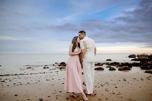 A couple in pink and white attire shares a romantic moment on a rocky beach during sunset.