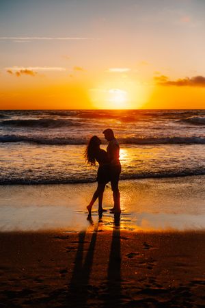 Silhouettes of a couple embrace on a beach at sunset as golden light reflects off ocean waves and sand.
