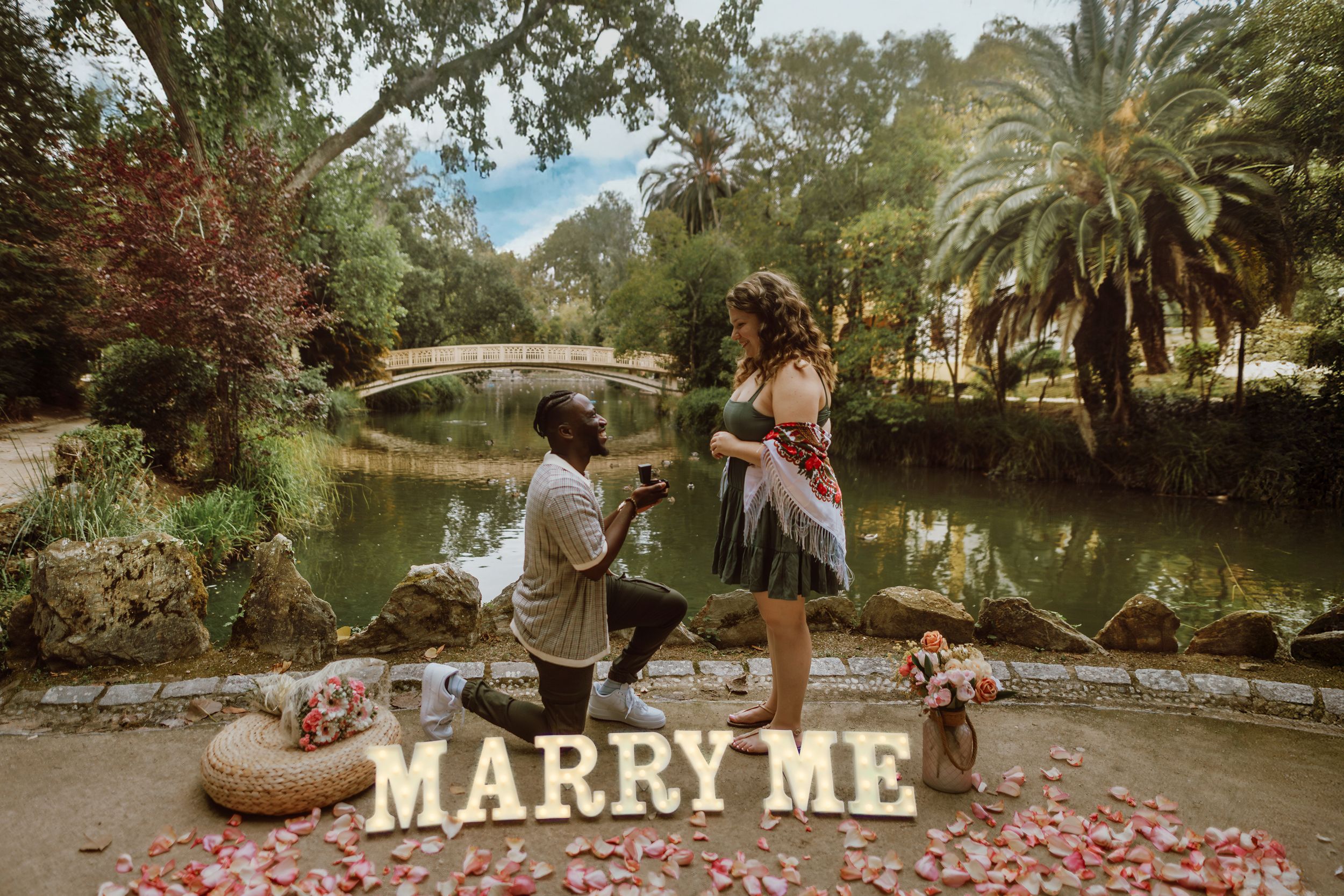 A romantic outdoor proposal setup with a lit 'MARRY ME' sign and rose petals scattered along a garden path at sunset.