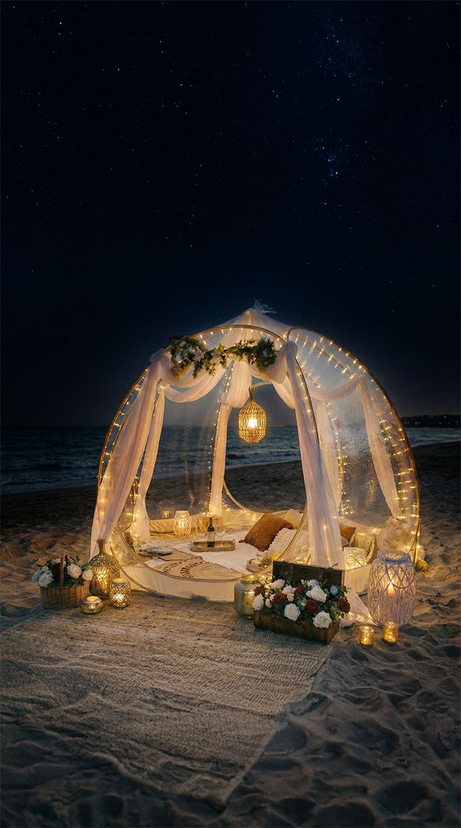 Romantic beach cabana with white drapes and twinkling lights creates an intimate dining setting under starlit sky.