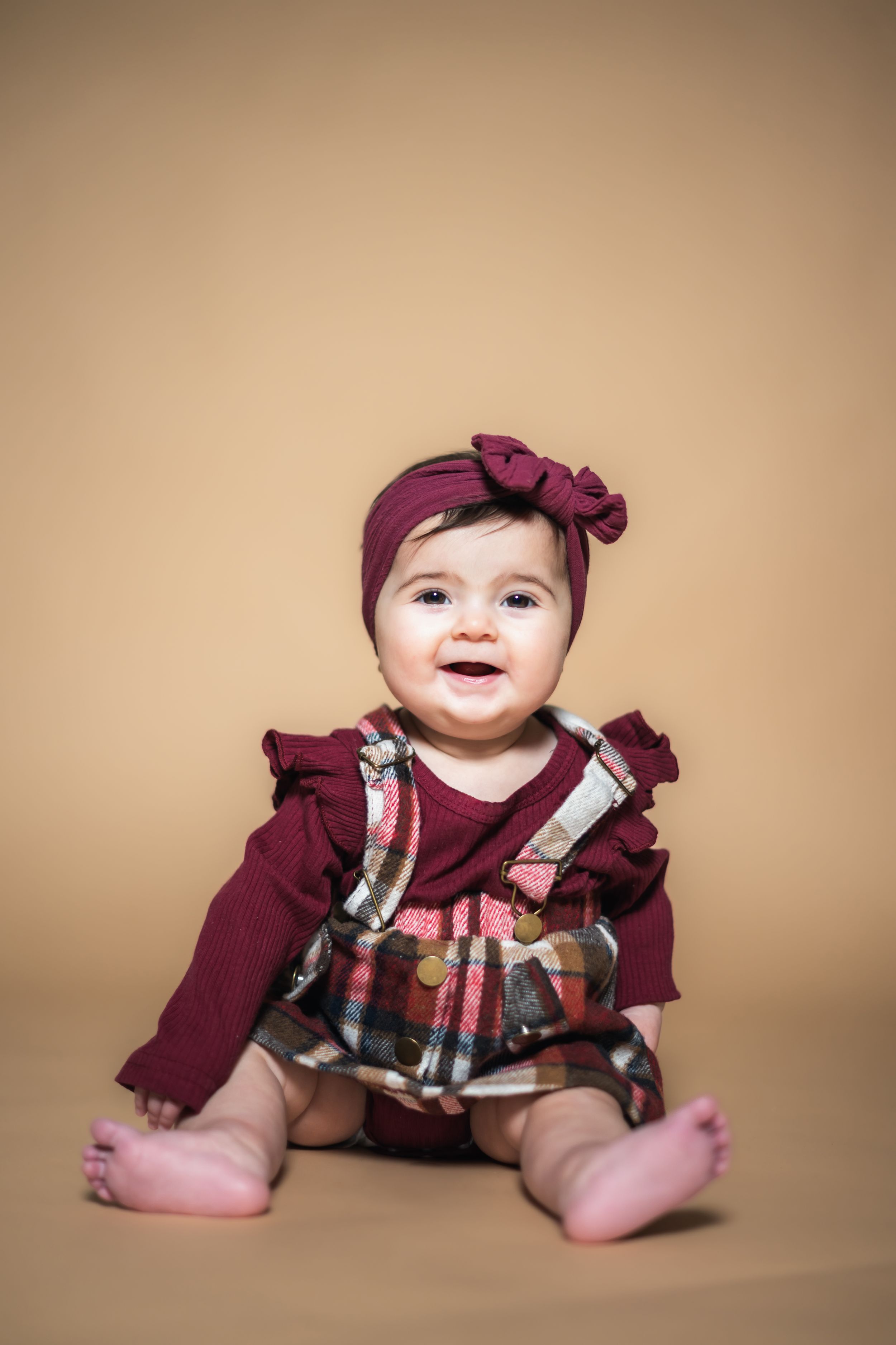 Smiling baby girl in burgundy ruffle top, plaid overalls, and matching bow headband sitting on beige background.