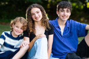 Three siblings sitting together outdoors wearing casual clothing against a blurred nature background.