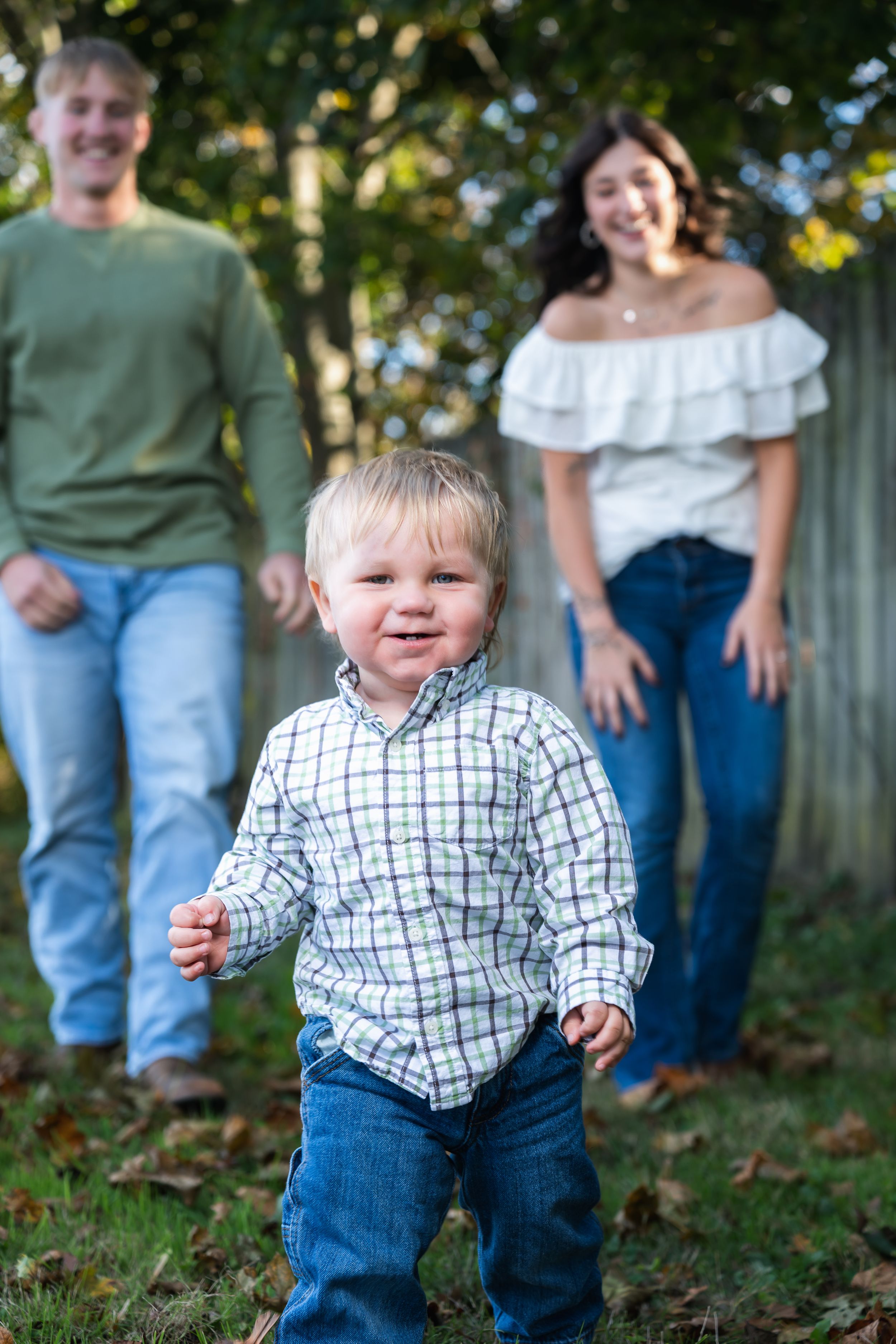 A happy toddler boy in a plaid shirt runs toward the camera while his smiling parents stand in a sunny backyard.
