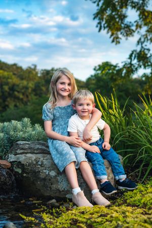 Two young siblings, a girl in a blue dress and a boy in white shirt and jeans, sit together on a rock by a pond.