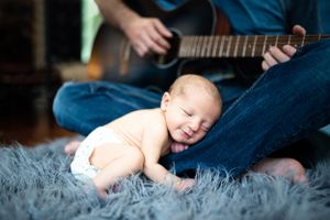 Baby in diaper laughing on gray fur rug while someone plays guitar.