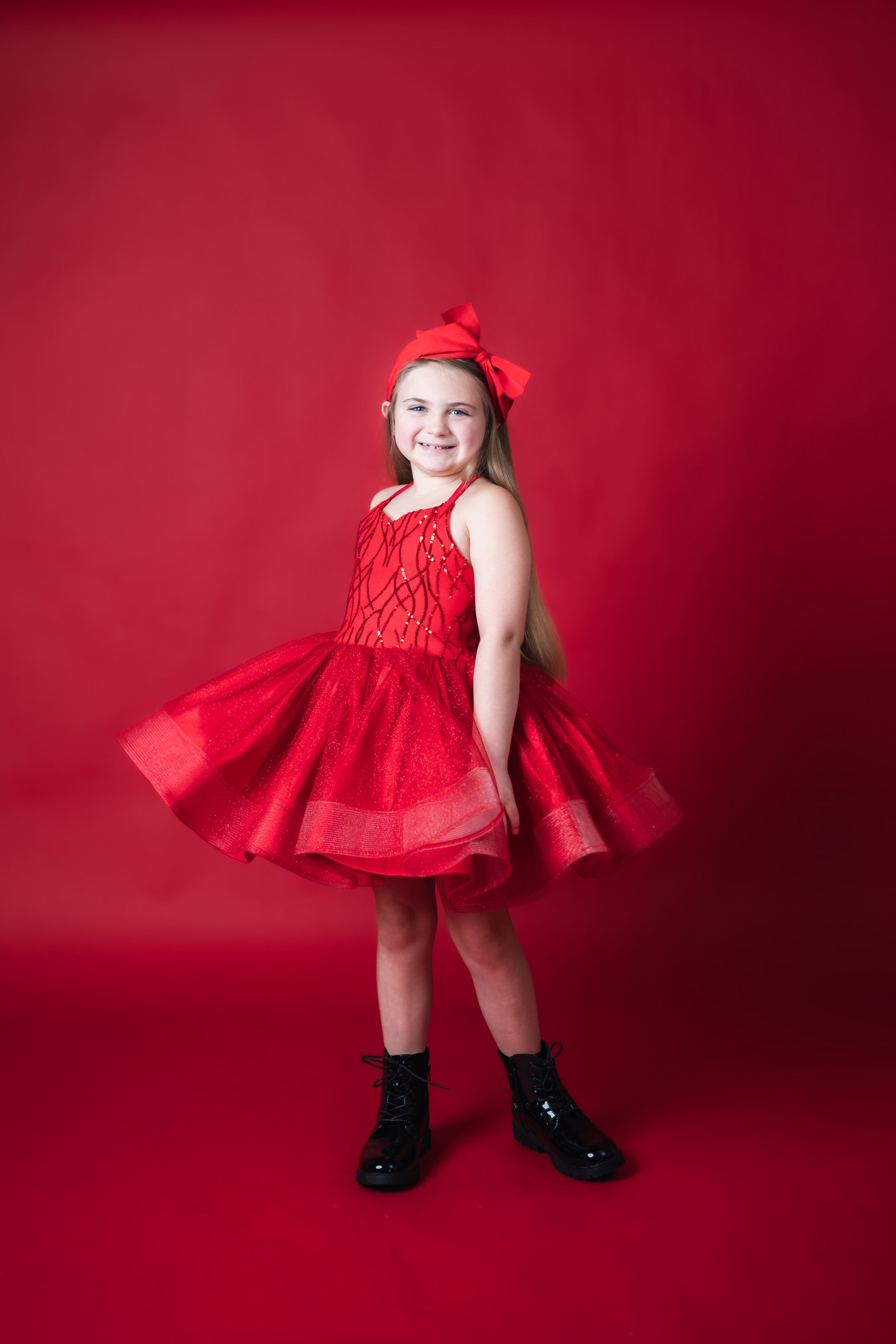 Young girl in red tulle dress and bow headband poses smiling against red background in black boots.