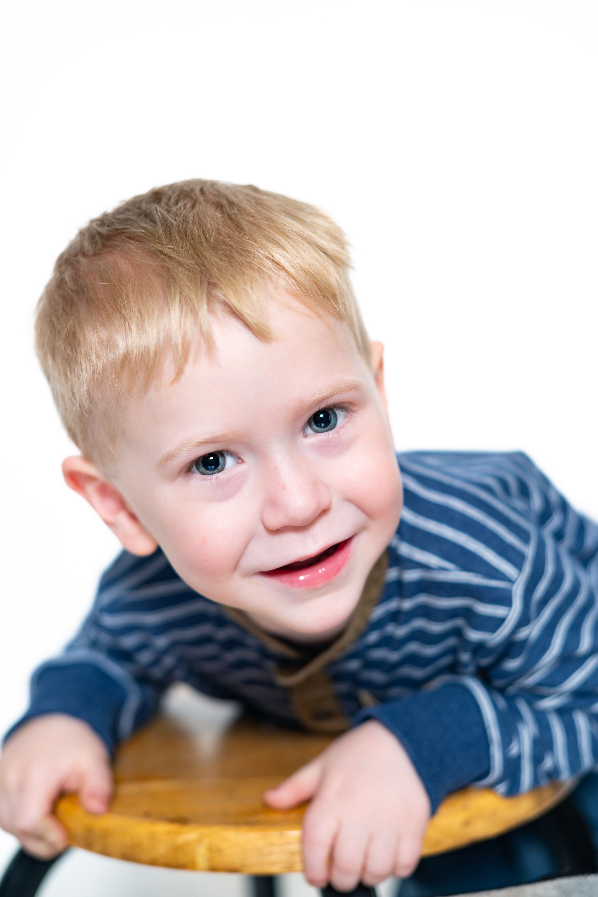 Young child in striped blue shirt leaning on wooden stool against white background.